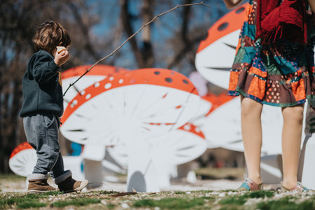 Child standing in a park next to oversized whimsical mushroom decorations holding a snack with an adult in colorful attire visible nearbyの写真素材
