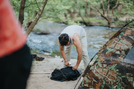 Man organizing backpack at a riverside campsite with surrounding natural sceneryの写真素材