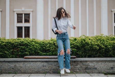 Confident young woman jumping outdoors wearing casual ripped jeans and sunglassesの写真素材