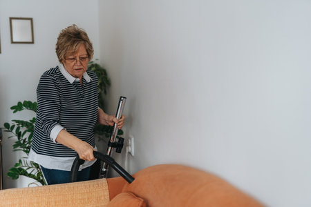 Senior Woman Vacuuming Living Room Beneath a Wall and Near a Houseplant, Focused on Keeping a Neat Homeの写真素材