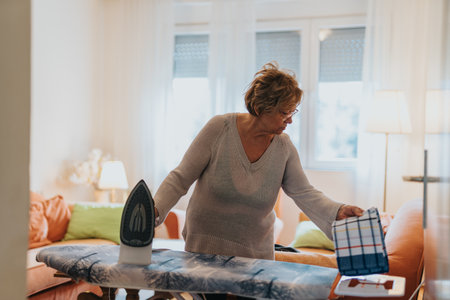 Senior woman ironing clothes in a bright cozy room with colorful decorの写真素材