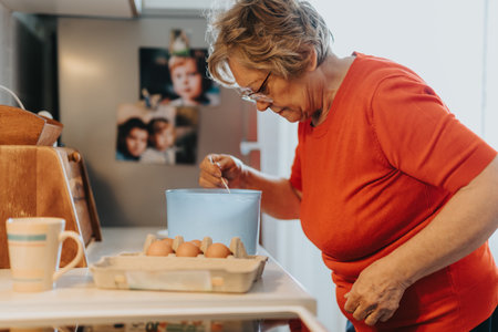 Elderly woman mixing ingredients in a blue bowl while cooking in a kitchen environmentの写真素材