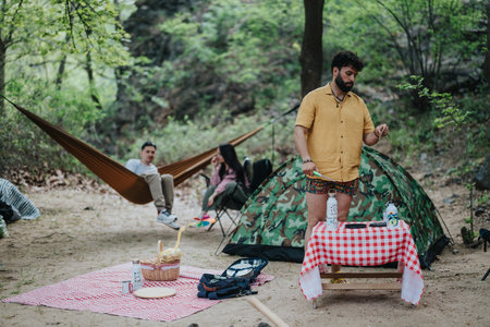 Friends camping outdoors with picnic setup and barbecue near a forest tentの写真素材