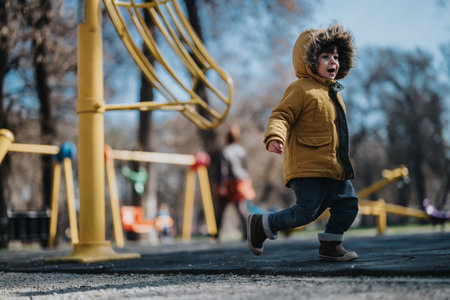 Young child in yellow winter coat enjoying playtime at a scenic outdoor park with vibrant playground equipment and trees.の写真素材