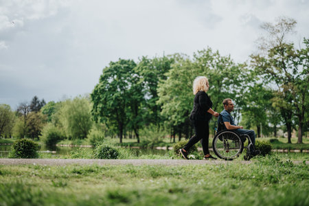 Woman assisting a man in a wheelchair along a park path surrounded by greenery and peaceful sceneryの写真素材