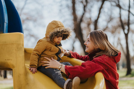 Mother and child enjoying playtime together on a playground slide on a cool day outdoors.の写真素材