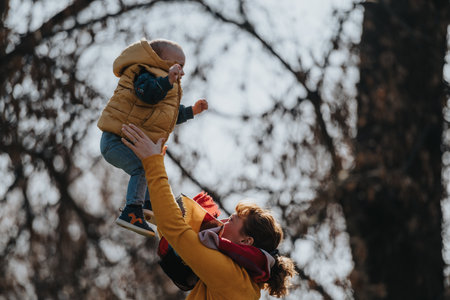 Mother lifts young child in a joyful play moment outdoors, expressing care and love on a sunny...