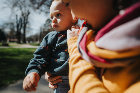 Close-up of a mother holding her child outdoors on a sunny day, with green grass and trees in the...