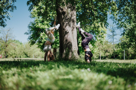 Two women exercising upside down against a tree in a sunny park for fitness and weight cutting.