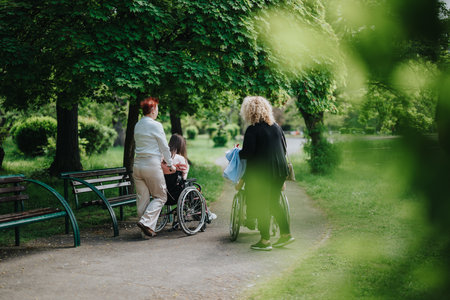 Back view of two women walking in a park, one assisting a child in a wheelchair.の写真素材