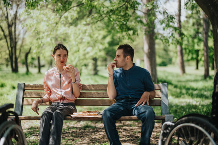 Young couple enjoys pizza while sitting together on a bench in a sunny park setting