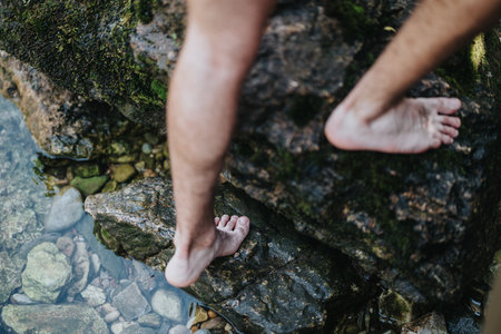Close-up of bare feet walking on mossy rocks beside a clear water streamの写真素材