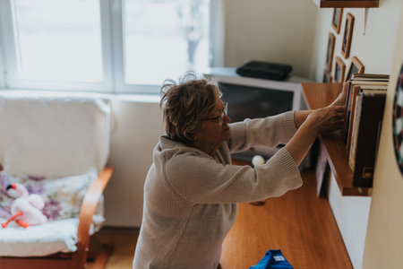Senior woman rearranging books on a shelf in a cozy living room interiorの写真素材