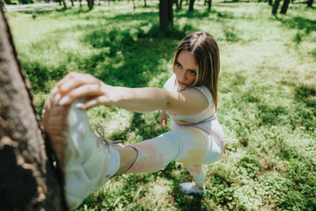 Woman stretching leg outdoors in park with trees and sunlightの写真素材