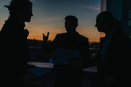 Business people are seen in silhouette, engaging in a thoughtful conversation against a vibrant sunset sky.の写真素材