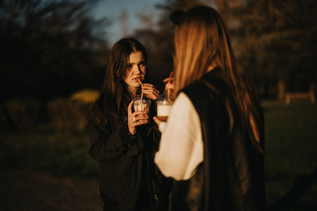 Two female teenagers enjoying drinks while having a conversation outdoors during a pleasant eveningの写真素材