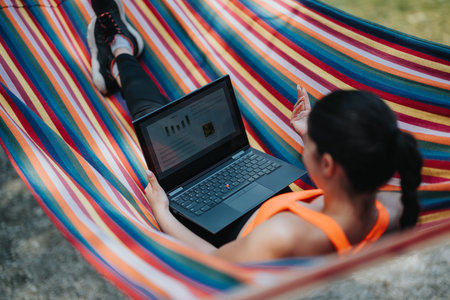 Woman Working Remotely While Relaxing on a Colorful Hammock Outdoorsの写真素材
