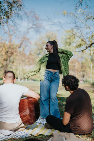A joyful outdoor gathering in a park with friends enjoying music and nature on a sunny day, featuring a woman dancing cheerfully among friends seated on a picnic blanket, creating a warm and lively atmosphere.の写真素材