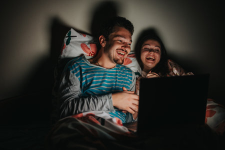 Happy couple watching a movie together in bed at nightの写真素材