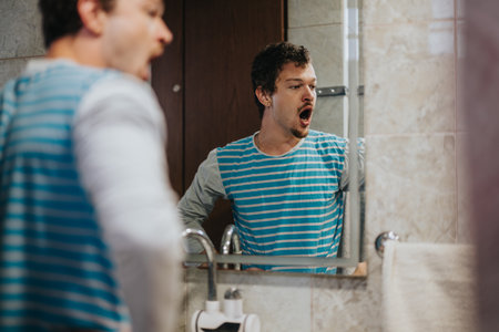 Young man yawning while looking at his reflection in a bathroom mirror, wearing a striped shirt and standing near a sink.の写真素材