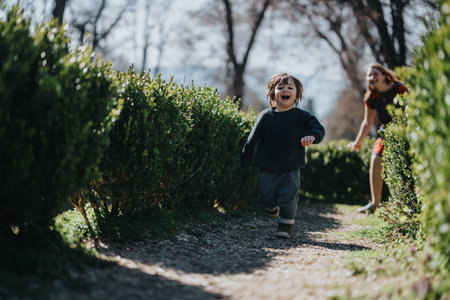 Delighted young child running through a green garden path with a parent following close behind on a sunny dayの写真素材