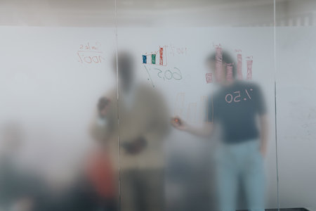Colleagues brainstorm on a project, writing data and charts on a glass board during a business...