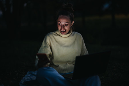 Young girl engrossed in phone and laptop during a nighttime settingの写真素材
