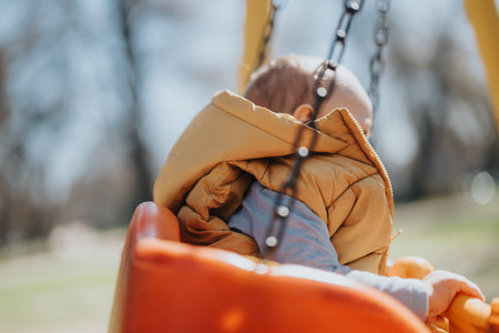 Young child wearing a yellow jacket enjoying time outdoors on a swing at a sunny parkの写真素材