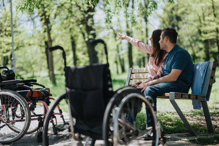 Couple enjoying nature on a bench, with wheelchairs nearby, within a park surrounded by vibrant green treesの写真素材