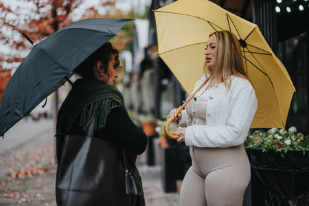 Two women with umbrellas chatting outdoors on an autumn dayの写真素材