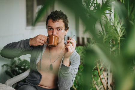 Young man drinking coffee and smoking cigarette surrounded by greenery at home outdoorsの写真素材