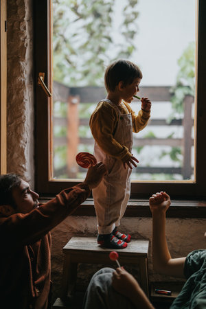 Child standing on a stool enjoying a lollipop while interacting with family indoorsの写真素材
