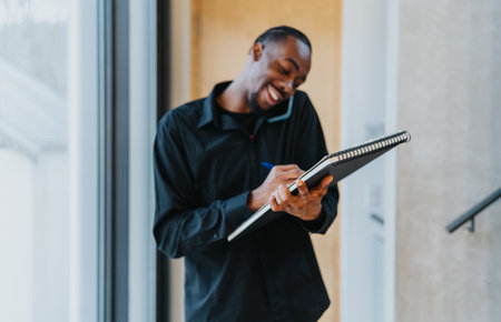 Smiling professional taking notes while engaging on a mobile phone conversation in a modern office settingの写真素材