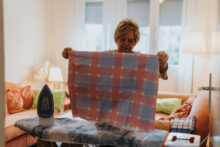 Elderly woman ironing plaid fabric in a cozy living room with natural lightの写真素材