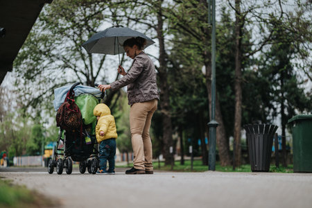 Mother with umbrella caring for child near stroller in a rainy parkの写真素材