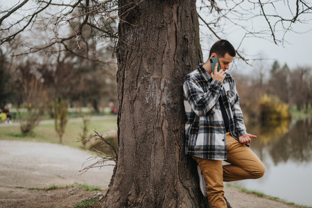 Man leans against tree while talking on phone in a calm park settingの写真素材