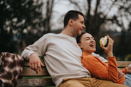 Happy couple relaxing on a park bench enjoying an outdoor snack togetherの写真素材