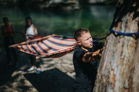 Man securing hammock outdoors near river with friends helping nearby