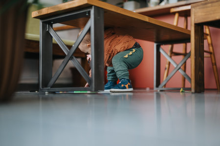Child crouching curiously beneath a wooden table in a cozy kitchen setting