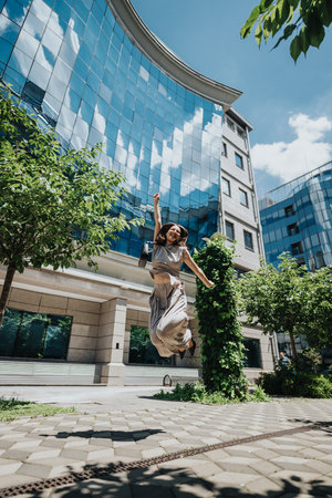 Young woman jumping joyfully outdoor near modern glass buildingの写真素材