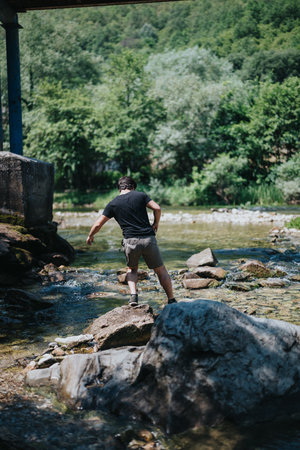 Man exploring river rocks beneath a bridge in a lush forest settingの写真素材