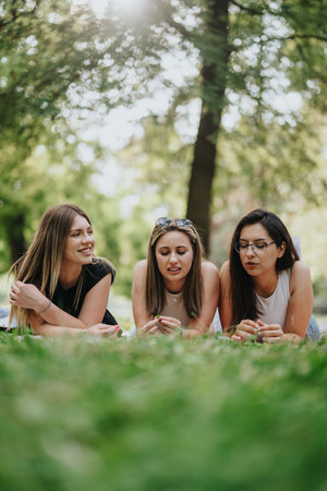Three young women enjoying the outdoors while relaxing on a sunny park lawnの写真素材