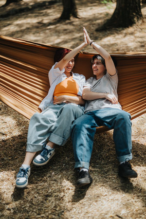 Friends share a playful high-five while relaxing in a hammock outdoors on a sunny dayの写真素材