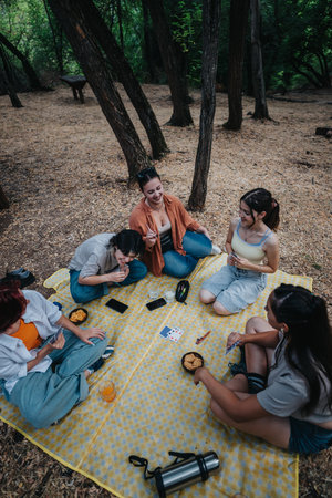 Friends enjoy a forest picnic and card game on a checkered blanketの写真素材