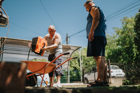 Two men work in a sunny yard loading a wheelbarrow with garden clippings