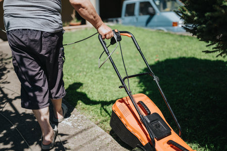 Person pushes orange lawn mower in sunny yard with green grass and a parked van in backgroundの写真素材