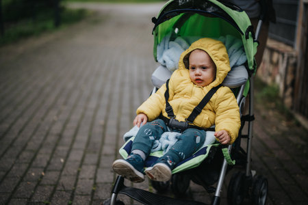 Child in cozy yellow jacket sitting in stroller on outdoor pathwayの写真素材