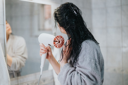 Woman drys wet hair with a hairdryer while wearing a robe at homeの写真素材