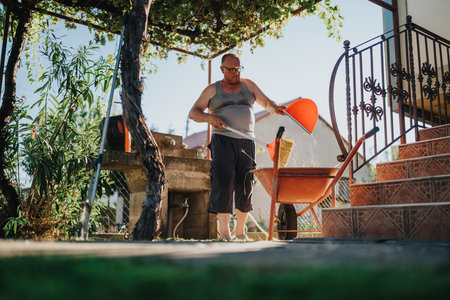 Man in sleeveless shirt cleaning yard with wheelbarrow, broom, and dustpan beside stairs and railingの写真素材