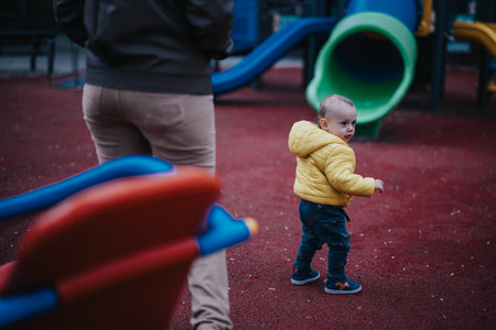 Toddler in a yellow jacket exploring playground with parent watching nearbyの写真素材